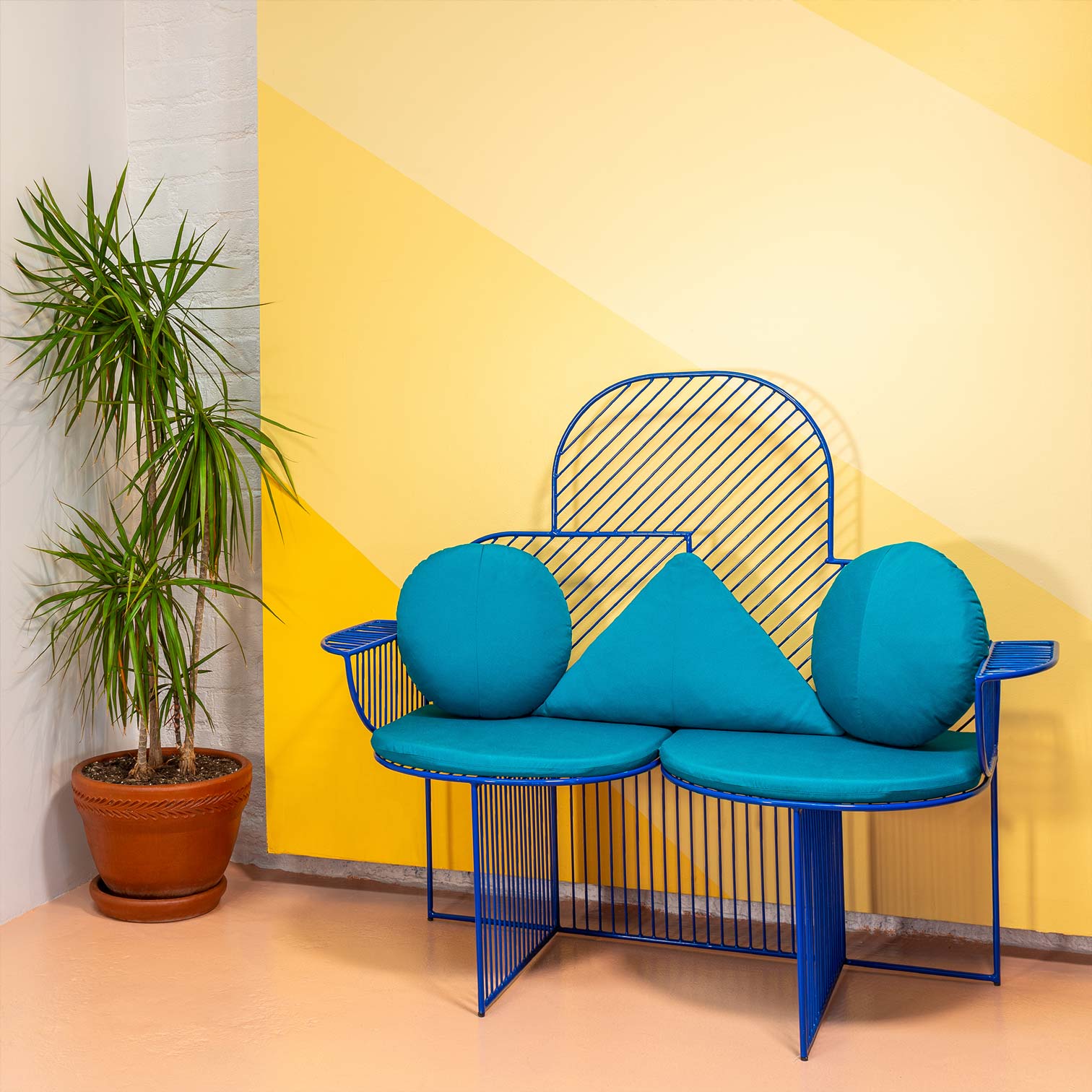 Cloud Bench shown in Electric Blue with Teal Sunbrella Padding, Triangle & Round Throw Pillows. Pictured in front of yellow art on a wall