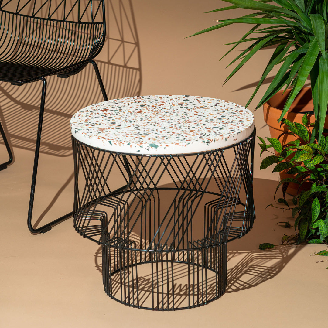 Terrazzo Table shown in Black. Pictured in front of tan backdrop, next to a stacking lucy chair and house plants.