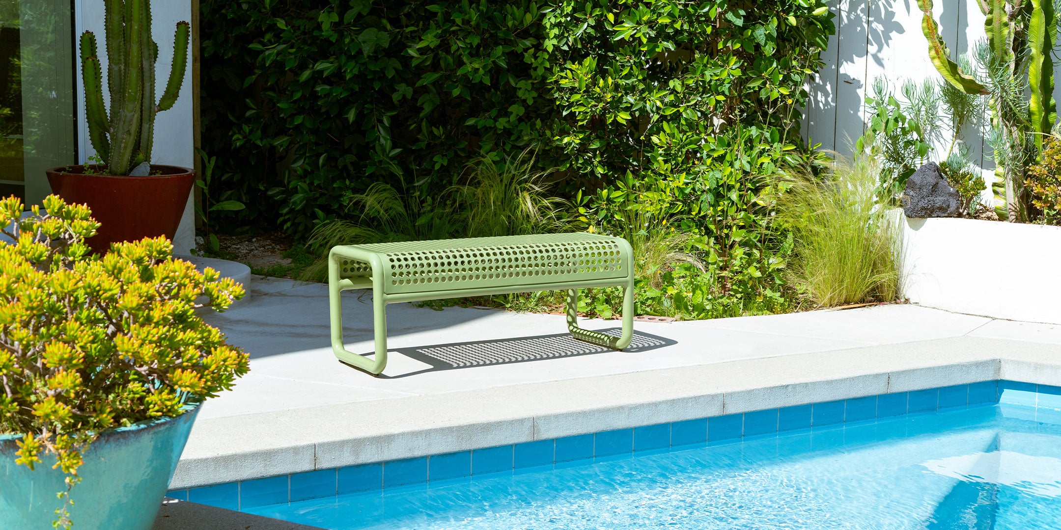 Green bench by a pool with plants and a cactus in the background. Tube Bench Indoor & Outdoor Perforated bench from Bend Goods