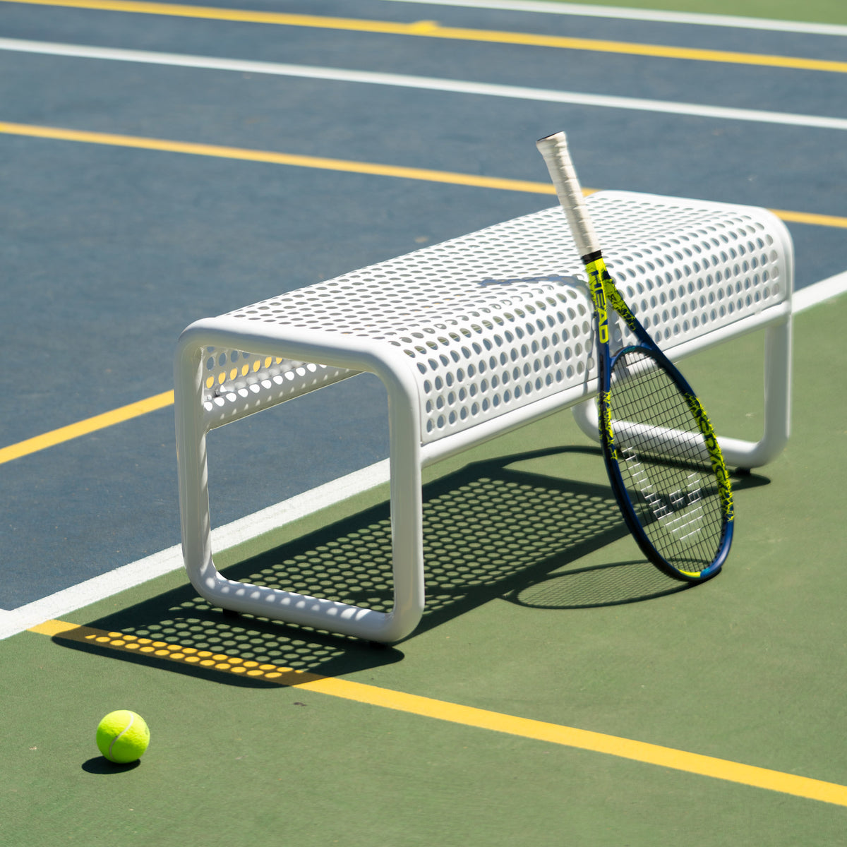 A white perforated metal tennis bench on an outdoor tennis court, with a tennis racket leaning against the back and a tennis ball on the court surface.